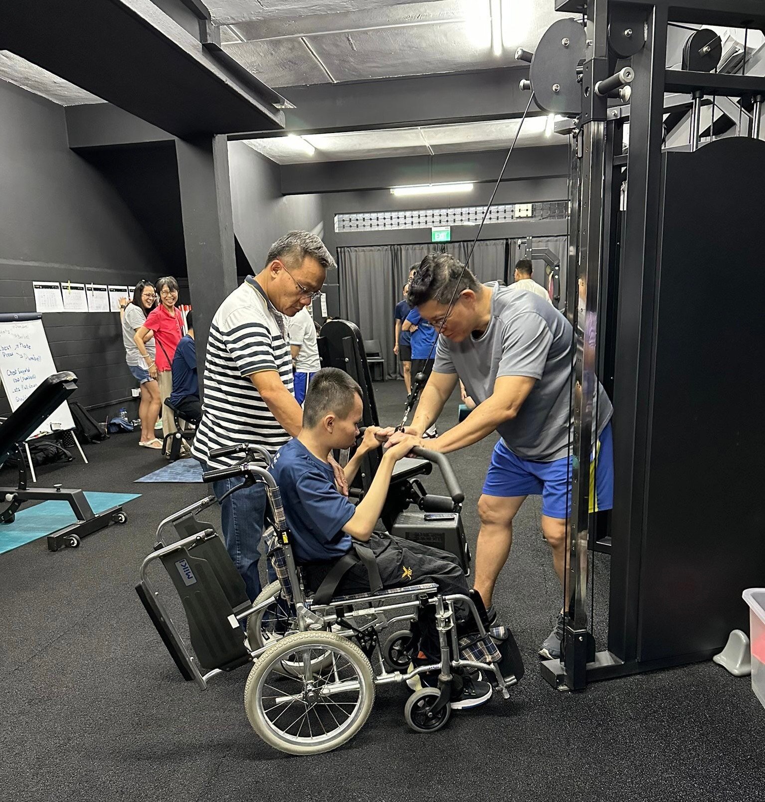 Two adults assisting a teenager on wheelchair using gym machine